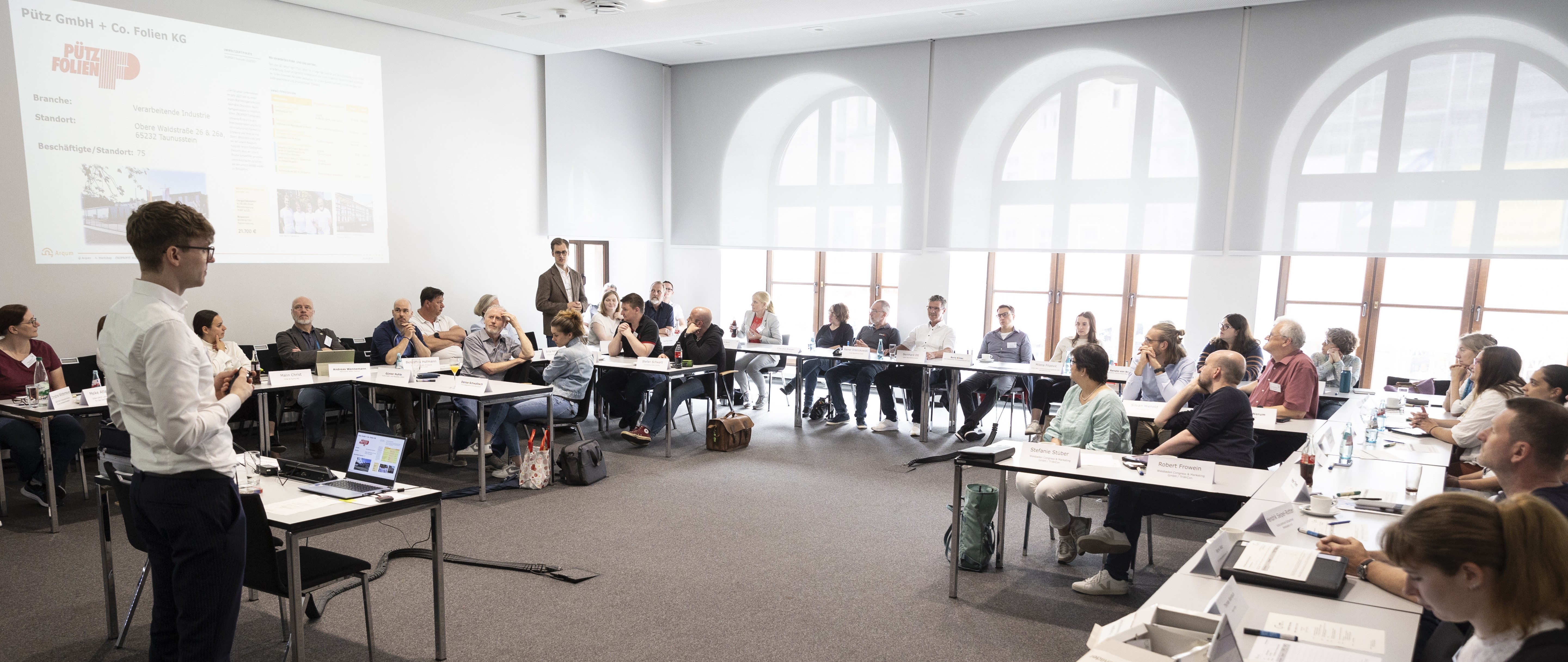 Workshop mit rund 30 Teilnehmenden in einem Konferenzraum. Eine Person präsentiert vor einer Leinwand, die Teilnehmenden sitzen an U-förmig angeordneten Tischen.
