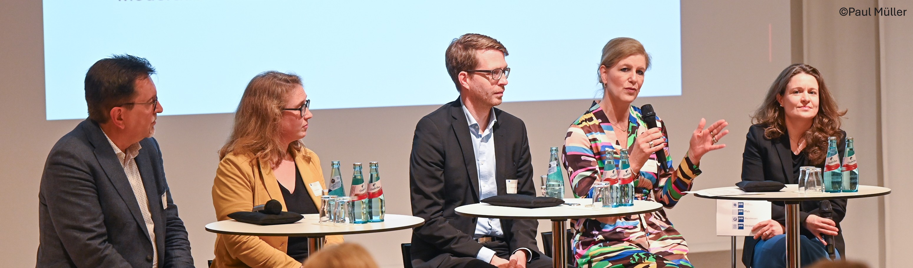 Podiumsdiskussion mit fünf Personen auf einer Bühne. Die Teilnehmenden sitzen an Stehtischen mit Mikrofonen und Wasserflaschen; eine Person spricht gerade ins Mikrofon vor einer Präsentationsleinwand.